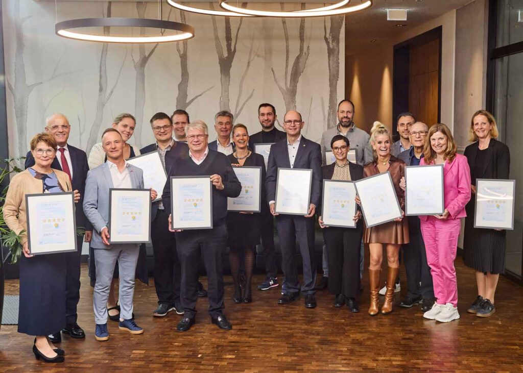 Eine Gruppe von Menschen steht in einem Raum, lächelt und hält gerahmte Zertifikate oder Auszeichnungen in der Hand. Sie sind in Geschäfts- und Freizeitkleidung gekleidet und posieren zusammen in einem modernen Raum mit Holzboden und dekorativer Wandkunst im Hintergrund.