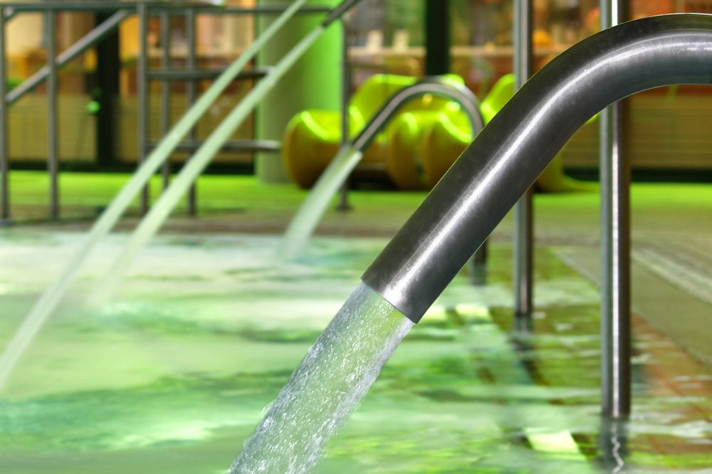 Ein Edelstahlrohr leitet Wasser in ein Hallenbad mit grüner Beleuchtung im „Wellness in Sole-Thermen“; im Hintergrund sind Wasserstrahlen und moderne gelbe Stühle zu sehen.