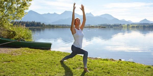 Eine Frau übt Yoga im Ausfallschritt mit erhobenen Armen im Freien auf grünem Gras neben einem ruhigen See. Umgeben von Bergen und Bäumen unter einem klaren Himmel, verkörpert sie den Geist der Wellness Stars Deutschland.