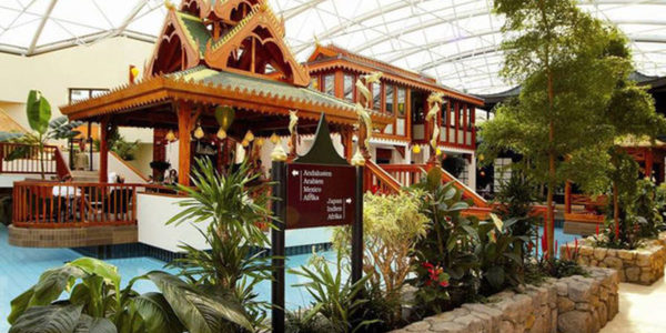 Innengarten mit tropischen Pflanzen und einem traditionellen Holzpavillon im thailändischen Stil, umgeben von einem kleinen blauen Pool unter einer Glaskuppel. Ein braunes Schild mit weißer Schrift im Vordergrund zeigt Wellness Stars Deutschland.