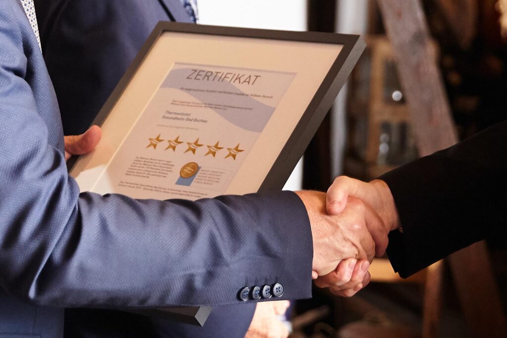 Eine Person im Anzug hält eine gerahmte Urkunde mit der Aufschrift Zertifikat und fünf goldenen Sternen in der Hand und schüttelt einer anderen die Hand - ein Hinweis auf Die Zertifizierung und das Prestige einer Preisverleihung oder Anerkennungsfeier.
