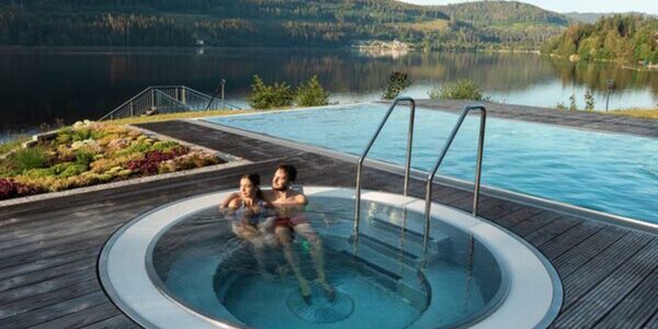 Ein Mann und Frau sitzen in einem runden Whirlpool im Außenbereich des Seehotels Wiesler. Im Hintergrund ist ein weiterer Pool sowie der See zu sehen.
