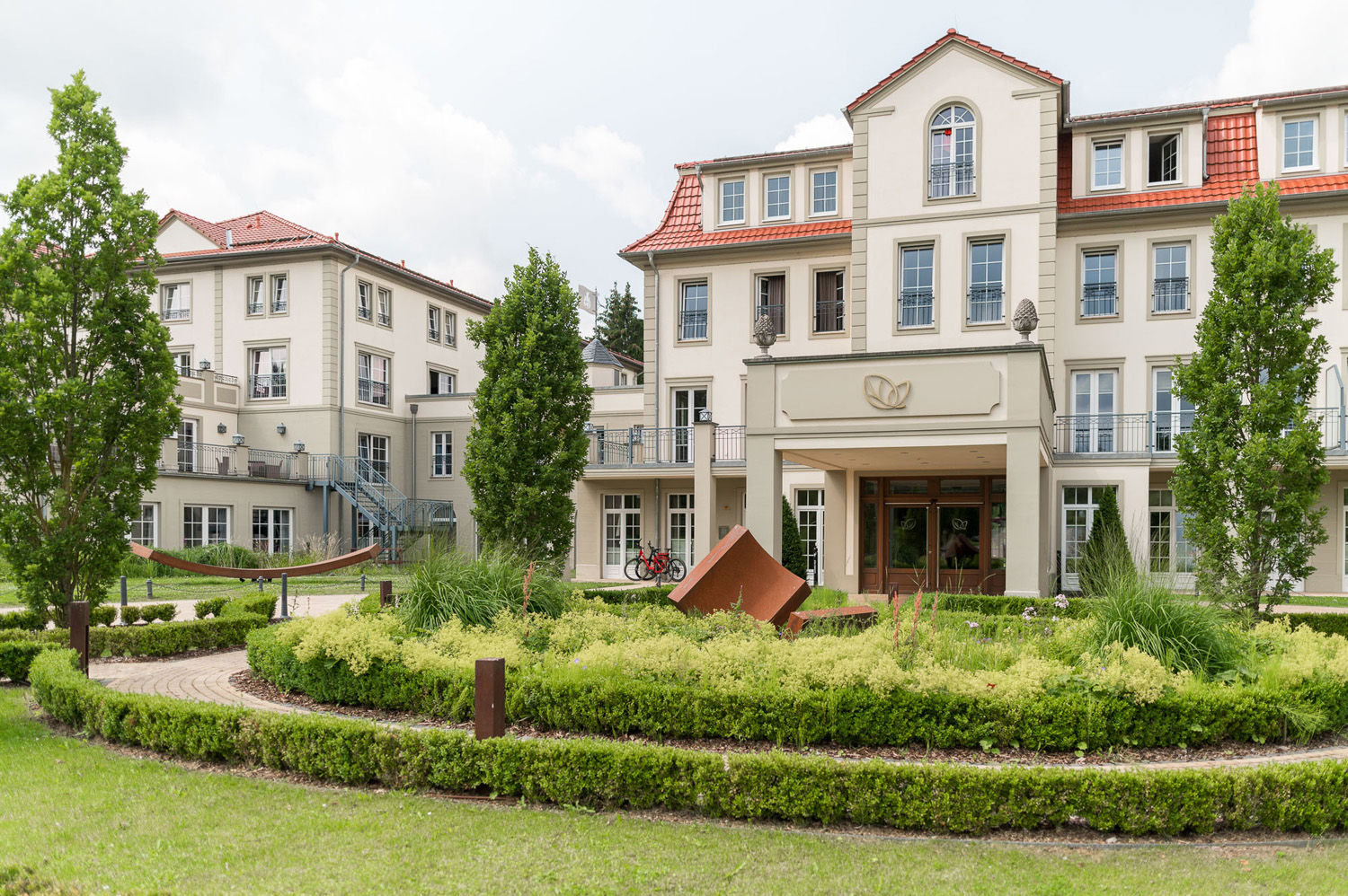 Ein großes, elegantes Gebäude mit roten Dachziegeln und vielen Fenstern, umgeben von gepflegter Landschaft, grünen Rasenflächen und Bäumen. Vor dem Eingang befindet sich eine runde Auffahrt mit einer modernen Skulptur.
