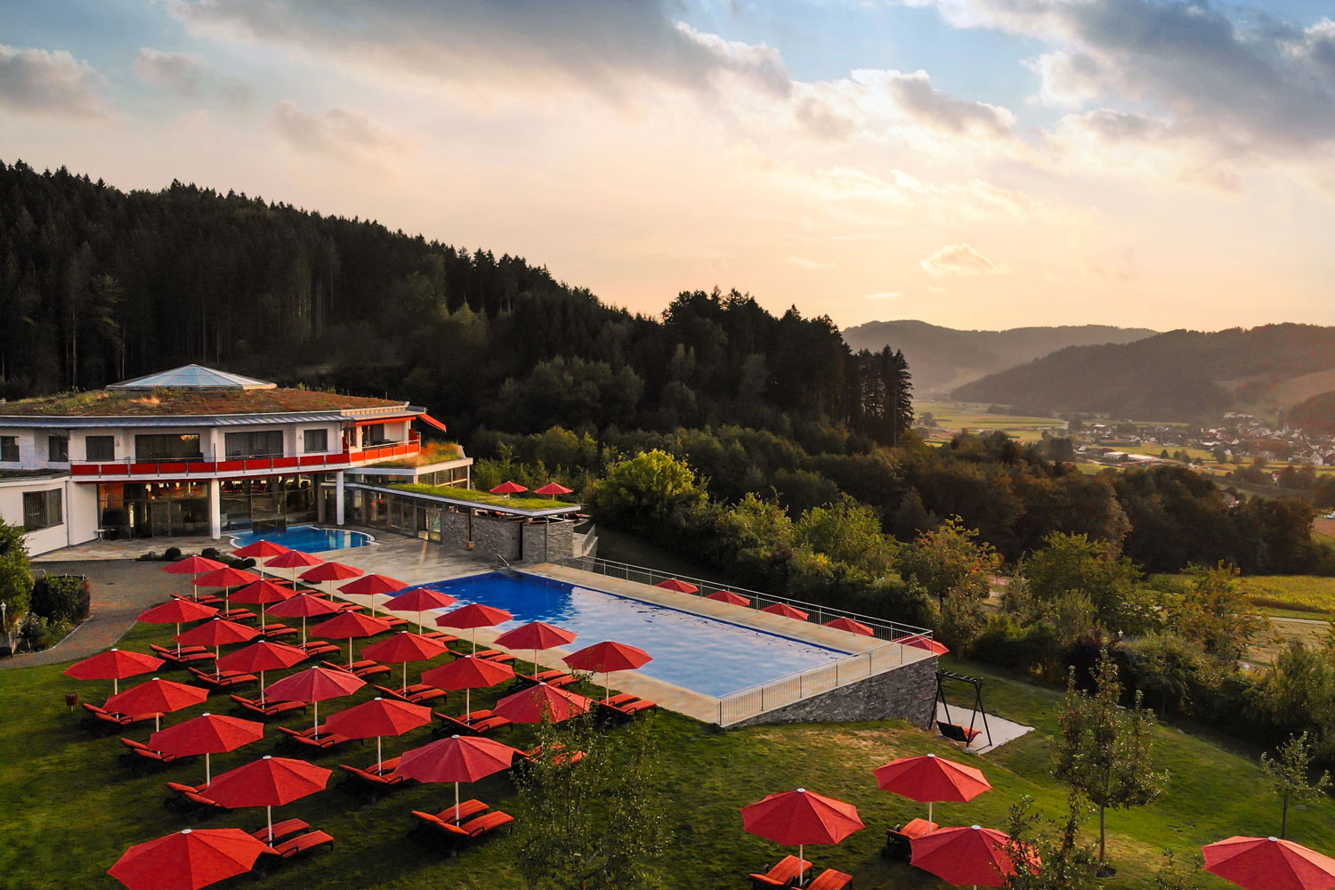 Ein landschaftlich reizvolles Resort mit einem großen Außenpool, umgeben von roten Liegestühlen und Sonnenschirmen, vor einer Kulisse aus Bäumen, sanften Hügeln und einem entfernten Dorf unter einem teilweise bewölkten Himmel bei Sonnenuntergang.
