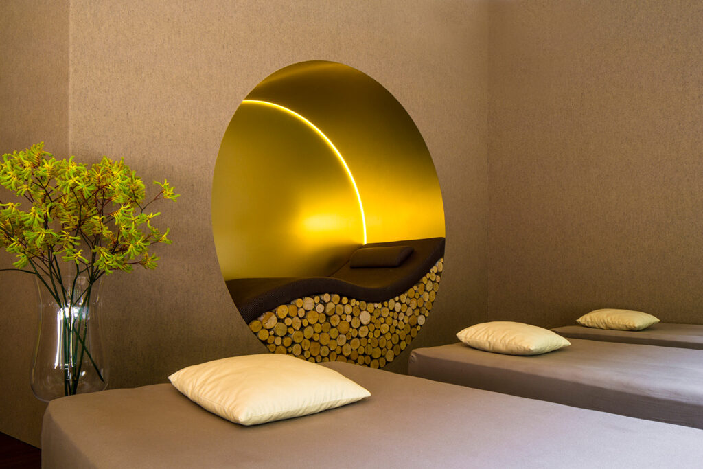Ein moderner Spa-Raum mit zwei Massagebetten, jedes mit einem weißen Kissen, und einer einzigartigen runden Nische mit gestapeltem Holz und goldener Beleuchtung. In der Nähe steht eine Glasvase mit gelb-grünen Blumen, die eine beruhigende Wirkung haben.