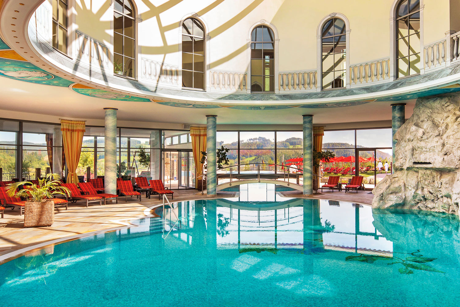 Luxuriöser Innenpool mit türkisfarbenem Wasser, umgeben von Liegestühlen, hohen Säulen und großen Fenstern, die eine herrliche Aussicht bieten. Das Sonnenlicht strömt durch eine Glaskuppel und schafft eine helle, einladende Atmosphäre.