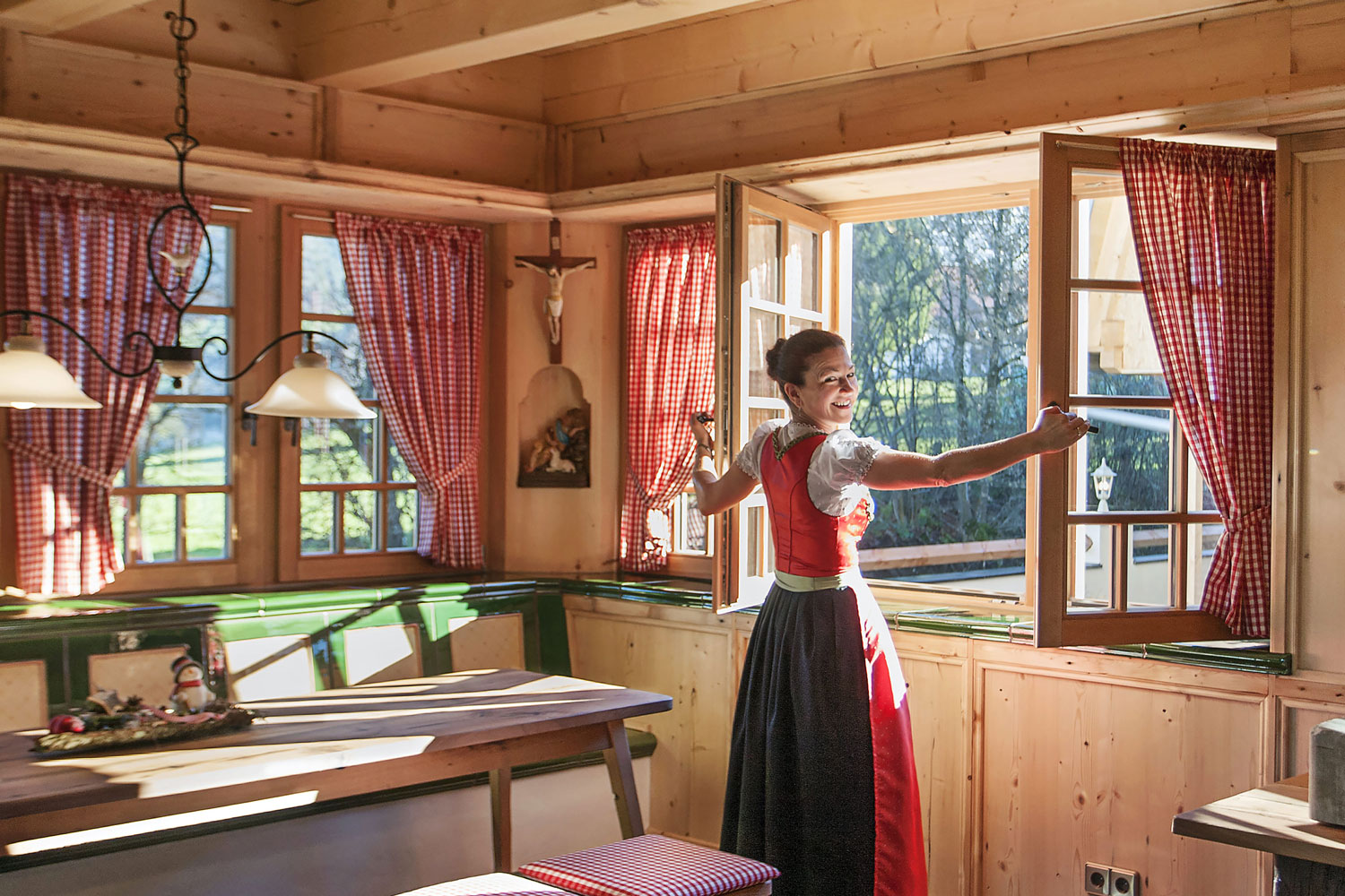 Eine Frau in bayerischer Tracht öffnet lächelnd ein Fenster in einer gemütlichen, sonnenbeschienenen Holzküche mit rotkarierten Vorhängen und rustikaler Einrichtung.