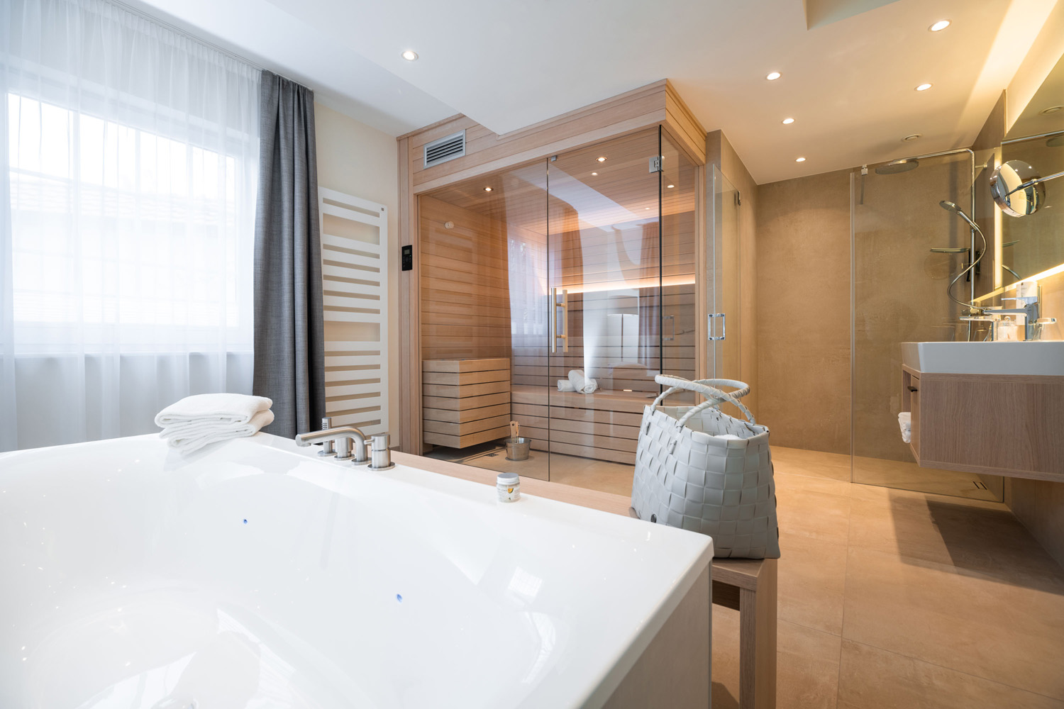 Modernes Badezimmer mit großer Badewanne, Weidenkorb, Doppelwaschbecken und einer verglasten Sauna; natürliches Licht strömt durch ein Fenster mit transparenten Vorhängen herein.