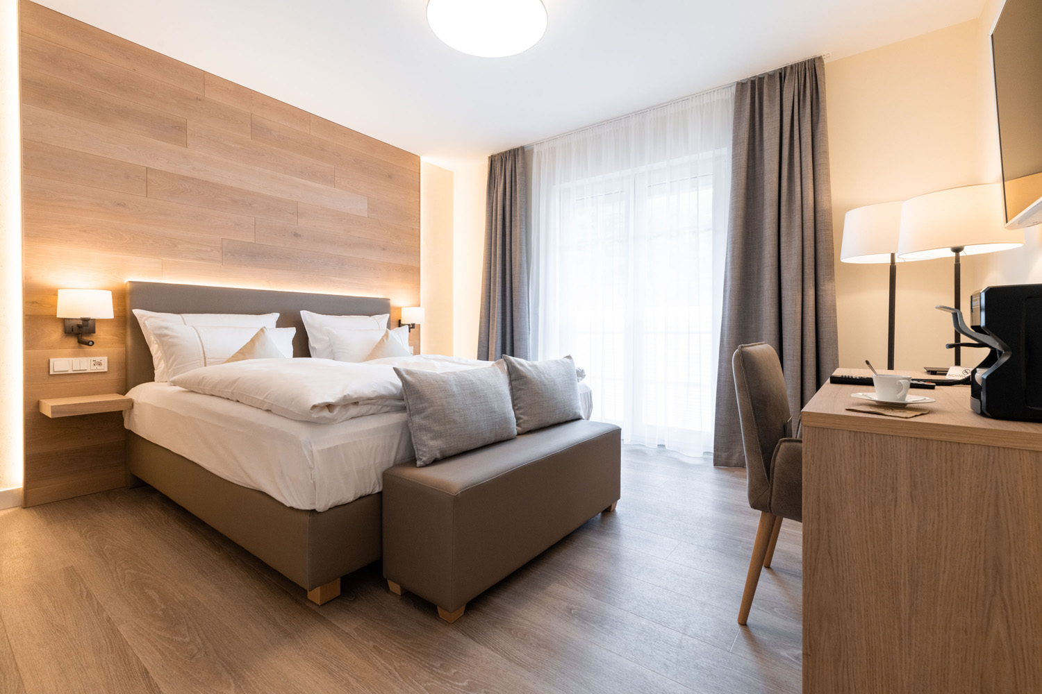 Ein modernes Hotelzimmer mit einem Doppelbett, grauer und weißer Bettwäsche, einer gepolsterten Bank am Fußende des Bettes, einem hölzernen Schreibtisch mit einem Stuhl, einer Stehlampe und einem großen Fenster mit durchsichtigen Vorhängen und grauen Gardinen.