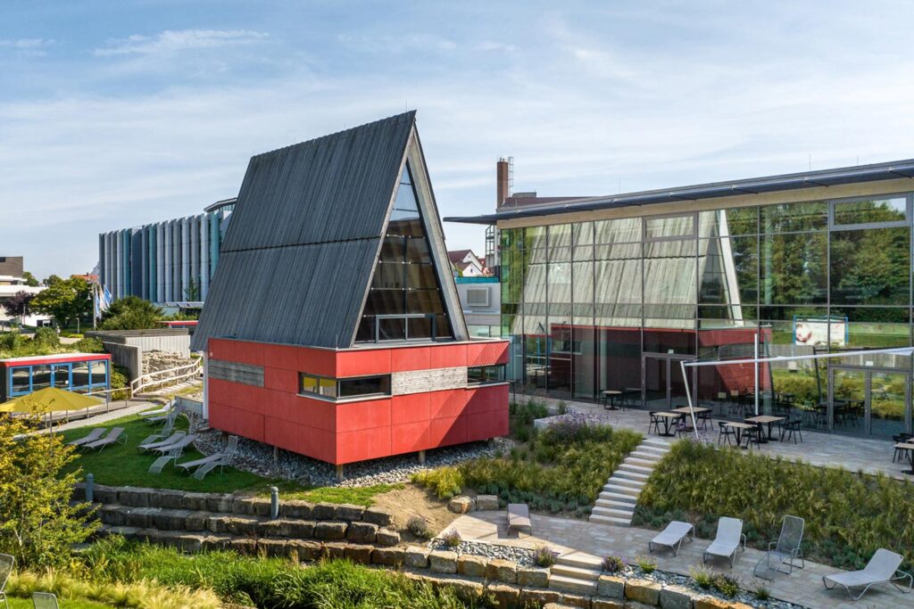 Ein modernes Gebäude mit einem steilen, dreieckigen A-Rahmen-Dach und roten unteren Wänden steht neben einem Glasbau. Es gibt Sitzgelegenheiten im Freien, Treppen, eine Grünanlage und ein weiteres buntes Gebäude im Hintergrund.