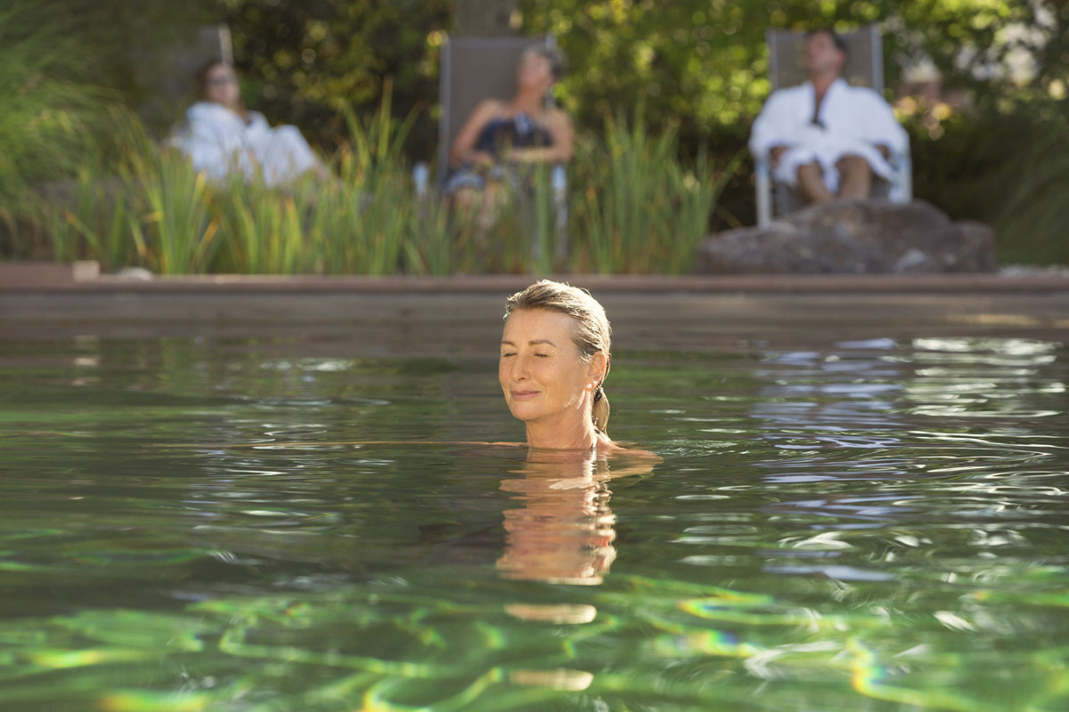 Eine Frau mit geschlossenen Augen entspannt sich in einem grün getönten Außenpool, während zwei Personen in Bademänteln auf Liegestühlen im Hintergrund sitzen, umgeben von üppigem Grün.