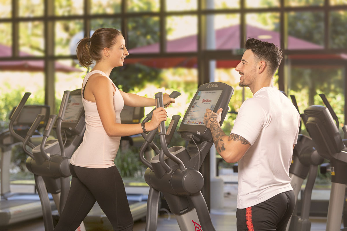 Eine Frau trainiert auf einer elliptischen Maschine, während ein Mann, möglicherweise ein Trainer, neben ihr steht und lächelt. Sie befinden sich in einem hellen Fitnessstudio mit großen Fenstern und mehreren Fitnessgeräten im Hintergrund.