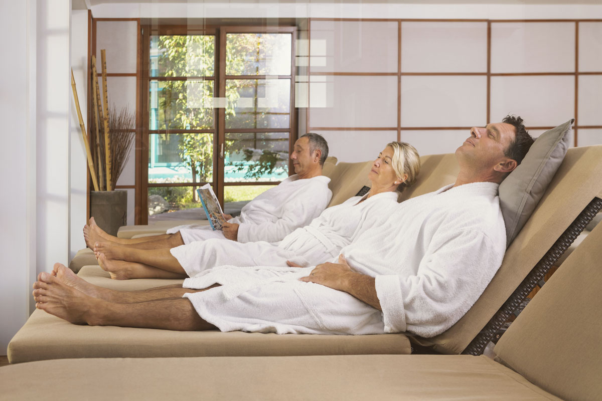 Drei Erwachsene in weißen Bademänteln entspannen sich auf Liegestühlen in einem hellen Wellness-Raum mit großen Fenstern und natürlichem Licht. Eine Person liest eine Zeitschrift, während die anderen mit geschlossenen Augen ruhen.