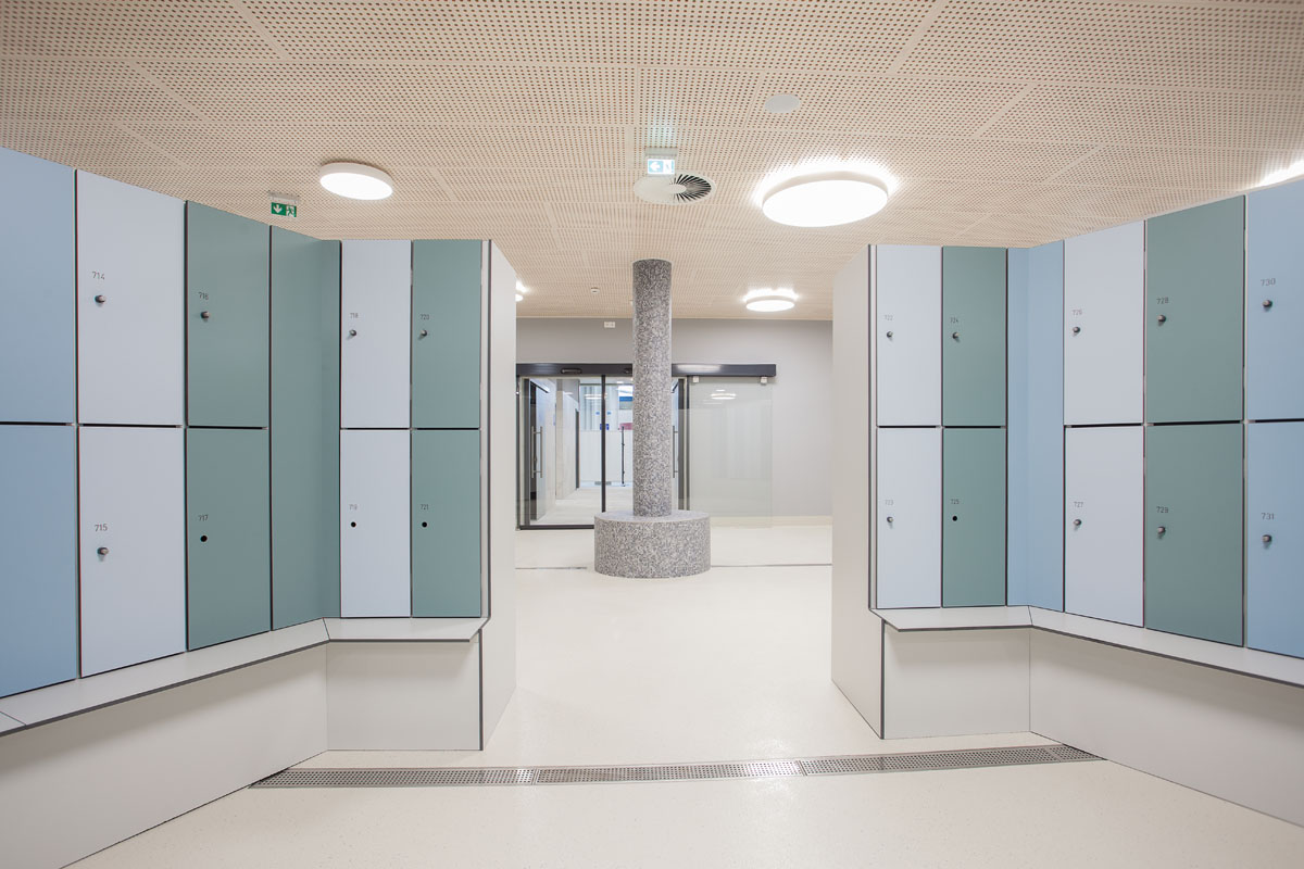 Ein moderner, sauberer Umkleideraum mit hellblauen und grauen Spinden auf beiden Seiten, heller Beleuchtung, einer runden Säule in der Mitte und Glastüren am anderen Ende.