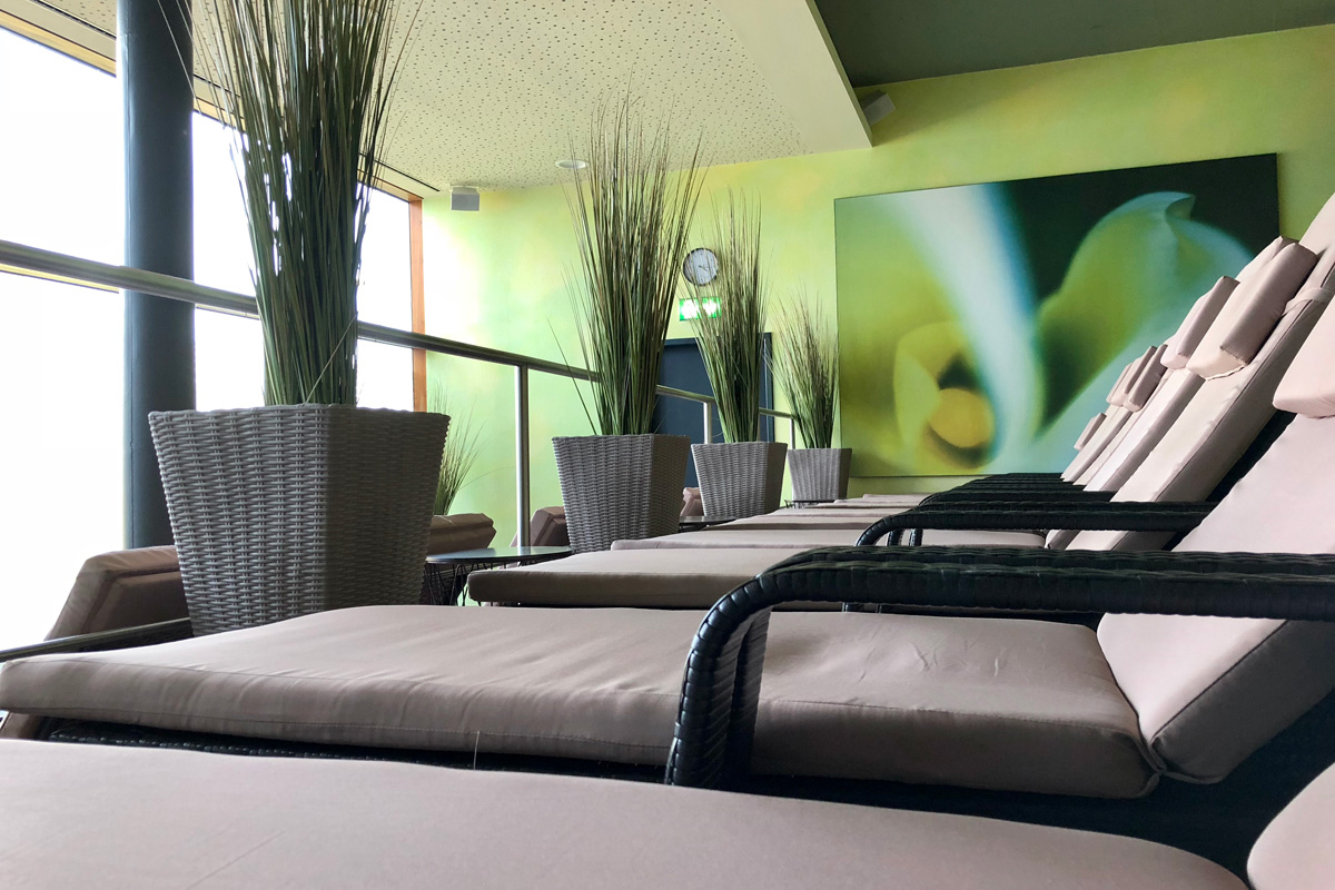 Eine Reihe von gepolsterten Loungesesseln mit dunklen Gestellen säumen einen Entspannungsbereich, der mit hohen Topfpflanzen und einem großen abstrakten Blumengemälde an einer grünen Wand dekoriert ist. Durch die großen Fenster fällt sanftes Tageslicht.
