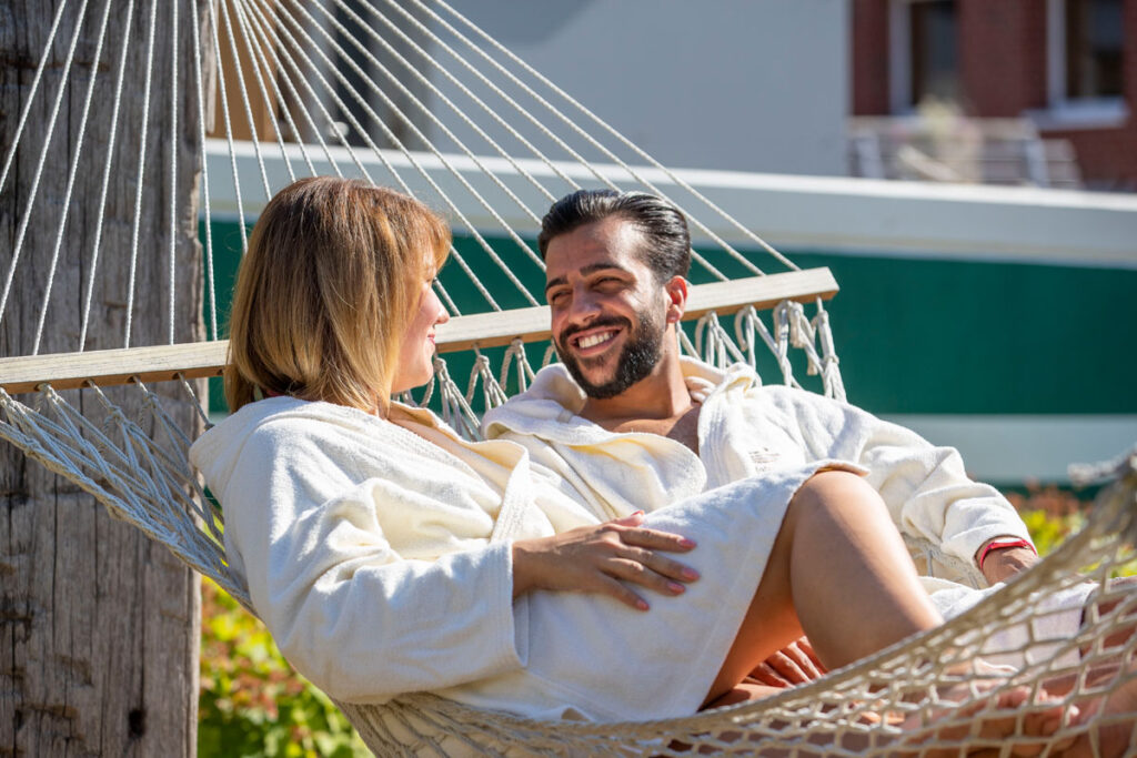 Ein Mann und eine Frau in weißen Gewändern entspannen sich gemeinsam in einer Hängematte im Freien, lächeln und genießen die Gesellschaft des anderen an einem sonnigen Tag.