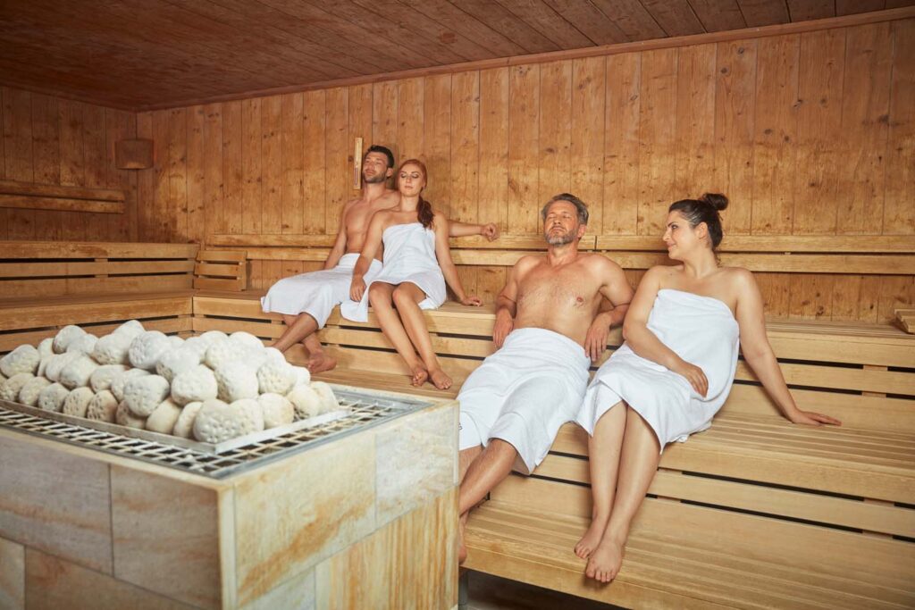 Vier Erwachsene in Handtüchern entspannen sich auf Holzbänken in einer Sauna. Die Sauna hat Holzwände und einen großen Haufen Saunasteine im Vordergrund.