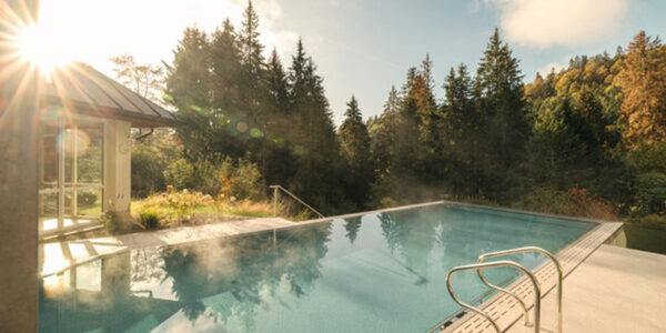 Ein ruhiger Außenpool in einem der Wellness-Hotels in Deutschland bietet einen Blick auf einen üppigen Wald mit hohen Bäumen, der in warmes Sonnenlicht getaucht ist. Wenn die Sonne durchscheint, steigt Dampf aus dem Wasser auf und erzeugt Lichtreflexe an einem Metallgeländer, das in den Pool führt.