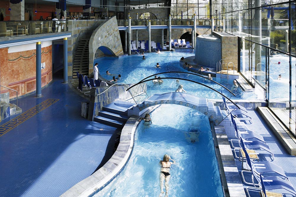 Hallenbadkomplex mit verschiedenen Schwimm- und Entspannungsbecken, darunter ein "Lazy River". Große Glasfenster lassen natürliches Licht herein, und am Beckenrand sind Liegestühle aufgestellt.