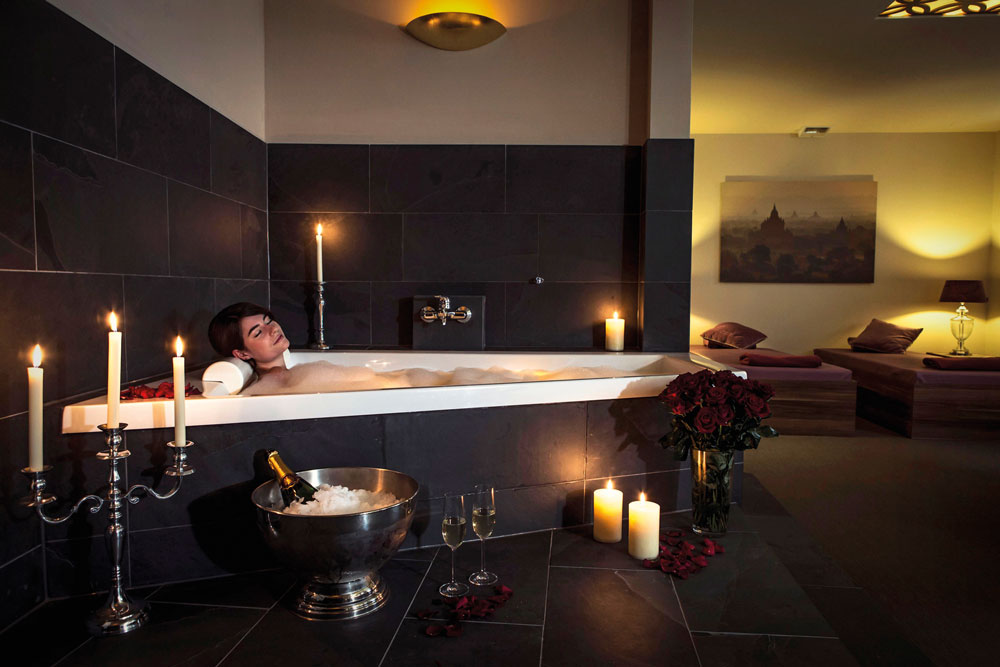 Eine Frau entspannt sich in einer schummrigen Badewanne, umgeben von Kerzen und Rosenblättern. In der Nähe stehen in einem Eimer eine Flasche Champagner und zwei Gläser, die eine luxuriöse und romantische Atmosphäre schaffen.