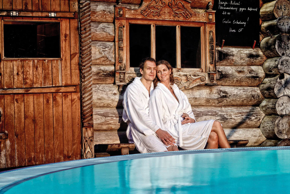 Ein Mann und eine Frau in weißen Gewändern sitzen eng beieinander auf einer Holzbank vor einer rustikalen Blockhütte und lächeln, während sich im Vordergrund ein Pool im blauen Himmel spiegelt.