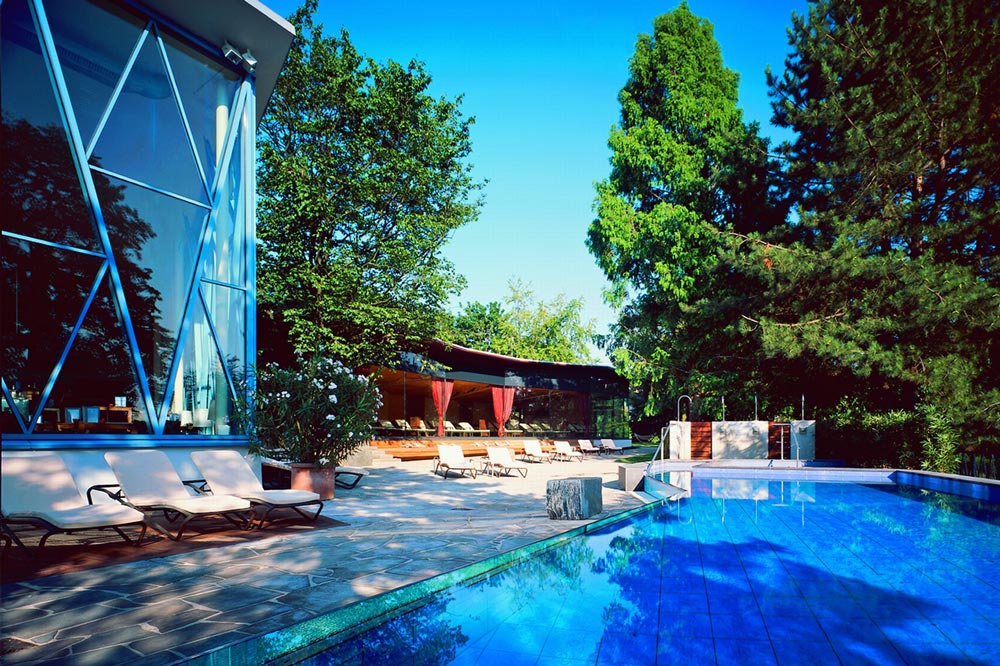 Außenschwimmbad mit klarem, blauem Wasser neben einem modernen Glasgebäude. Liegestühle und Sonnenliegen säumen die Steinterrasse unter grünen Bäumen. Der Bereich ist hell und sonnig, umgeben von üppiger Vegetation.
