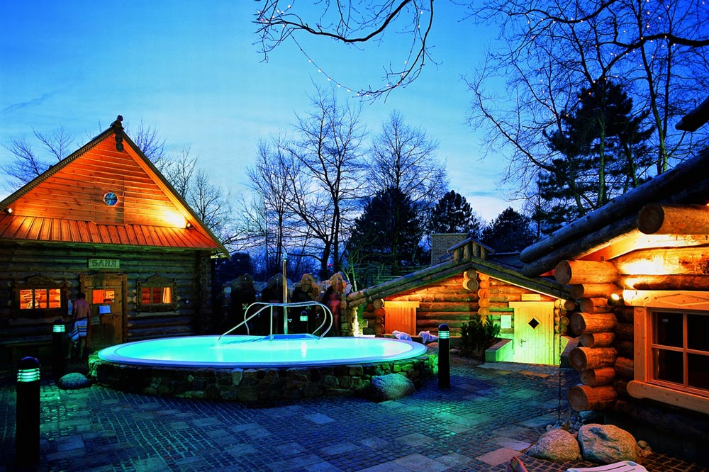 Ein runder, beleuchteter Außenpool ist in der Abenddämmerung von hölzernen Blockhütten umgeben, mit kahlen Bäumen und einem blauen Himmel im Hintergrund. Warme Lichter leuchten aus den Hütten und schaffen eine gemütliche Atmosphäre.