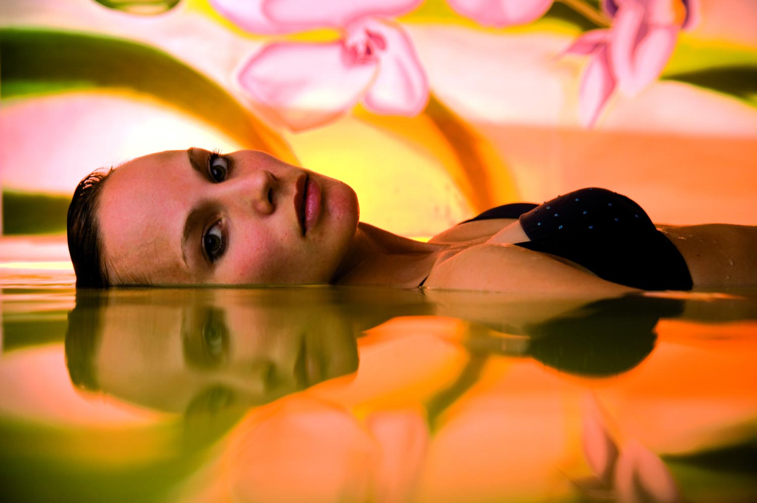 Eine Frau im schwarzen Badeanzug schwimmt auf dem Wasser, ihr Gesicht teilweise über der Wasseroberfläche, mit einer bunten Wandmalerei aus rosa Blumen und grün-gelben Wirbeln im Hintergrund, und ihr Spiegelbild ist im Wasser sichtbar.