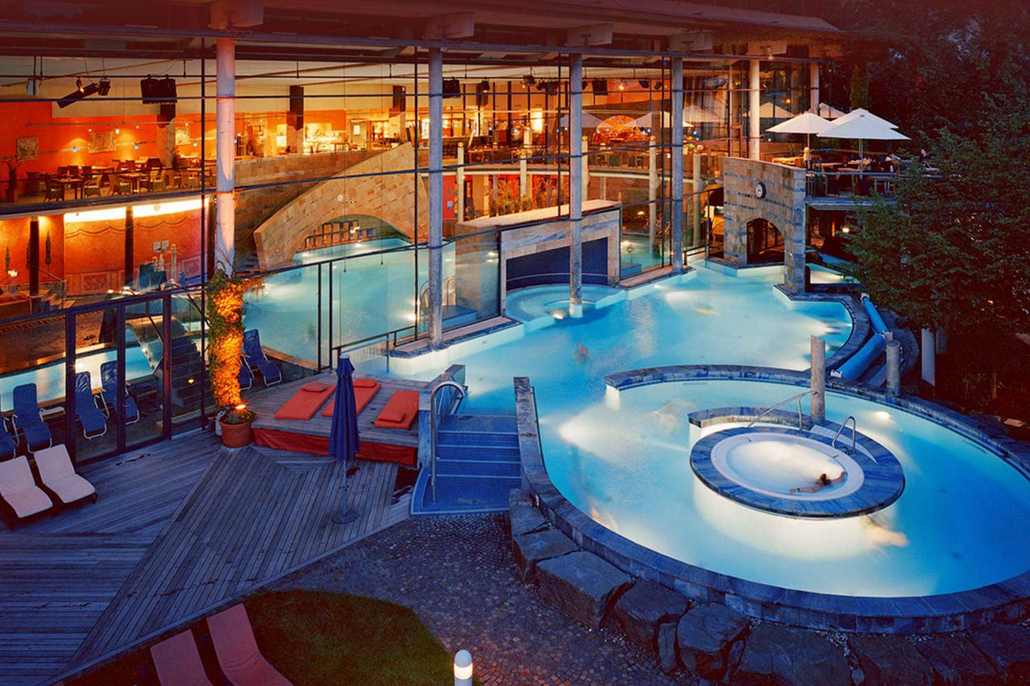 Ein nächtlicher Thermalbadkomplex im Freien mit einem runden Whirlpool, Liegestühlen, roten Liegen und einem Innenbereich mit großen Fenstern und stimmungsvoller Beleuchtung. Die Menschen entspannen sich im warmen, blau beleuchteten Wasser.