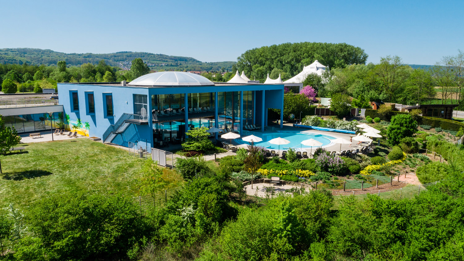 Ein modernes blaues Gebäude mit großen Glasfenstern steht neben einem Außenpool, der von Liegestühlen und Sonnenschirmen umgeben ist, inmitten von üppigem Grün und Gärten mit Hügeln und Bäumen im Hintergrund.