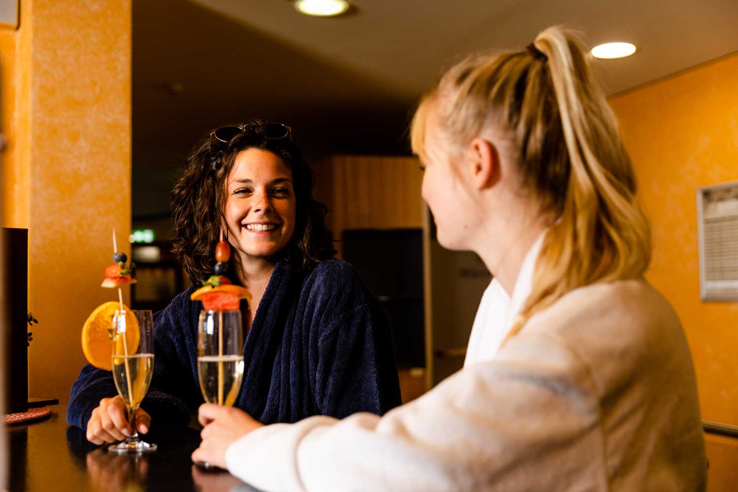 Zwei Frauen lächeln und unterhalten sich an einer Bar, jede hält ein mit Obst garniertes Cocktailglas. Beide tragen legere, bequeme Kleidung, und der Hintergrund ist in warmen, gelben Tönen gehalten.