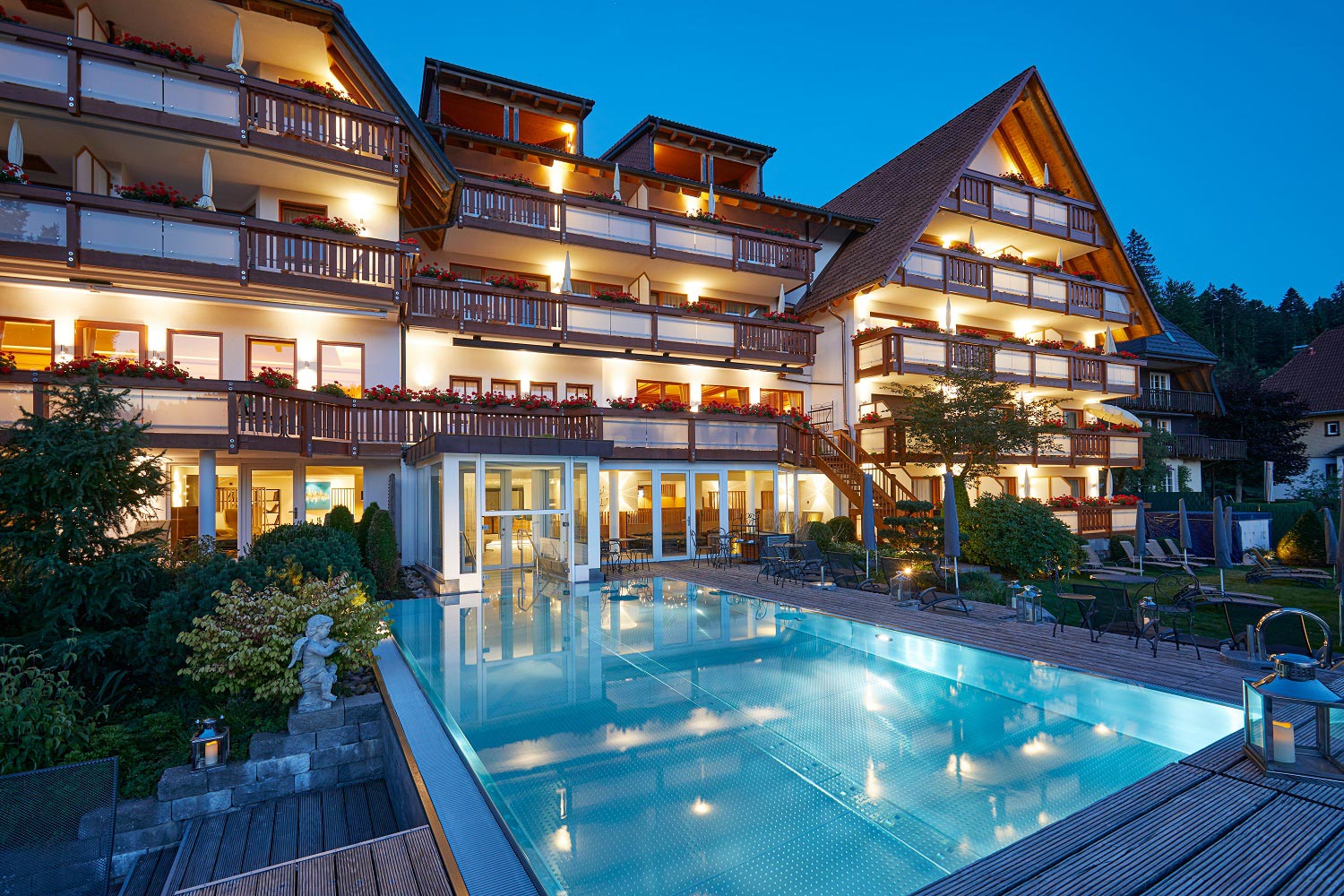 Ein großes, elegantes Hotel mit hölzernen Balkonen und leuchtenden Lichtern überblickt in der Abenddämmerung einen klaren Swimmingpool im Freien, umgeben von Bäumen und angelegten Gärten.