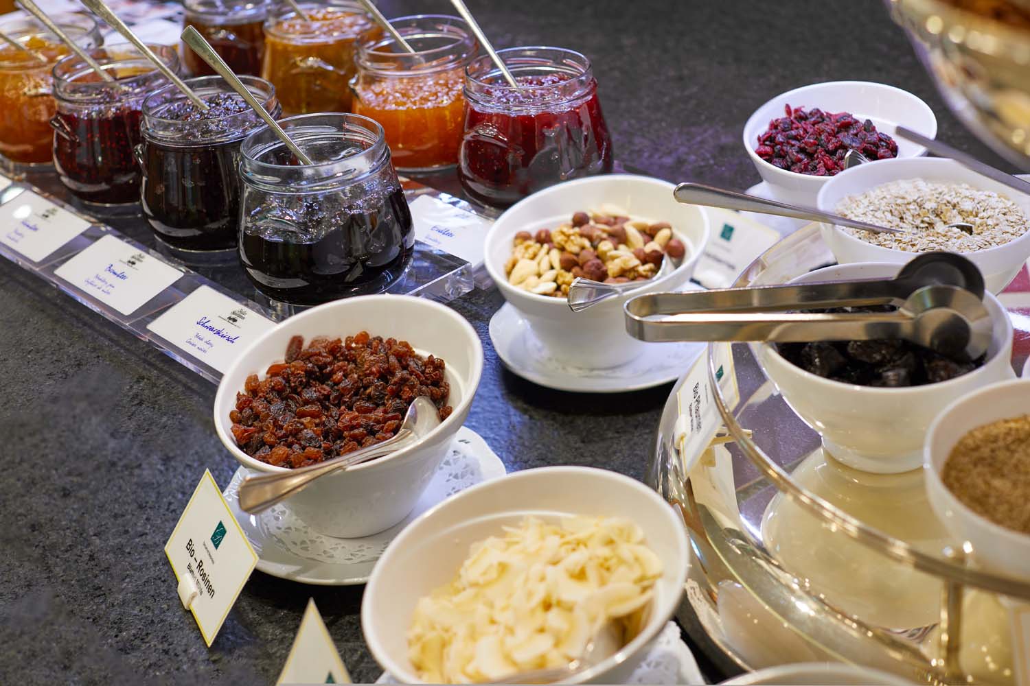 Ein Frühstücksbuffet mit Schalen mit Trockenfrüchten, Nüssen und Gläsern mit verschiedenen Marmeladen, jeweils mit einem Löffel, auf einer Arbeitsplatte mit Etiketten. Auf einigen Schalen liegt eine Zange zum Servieren.