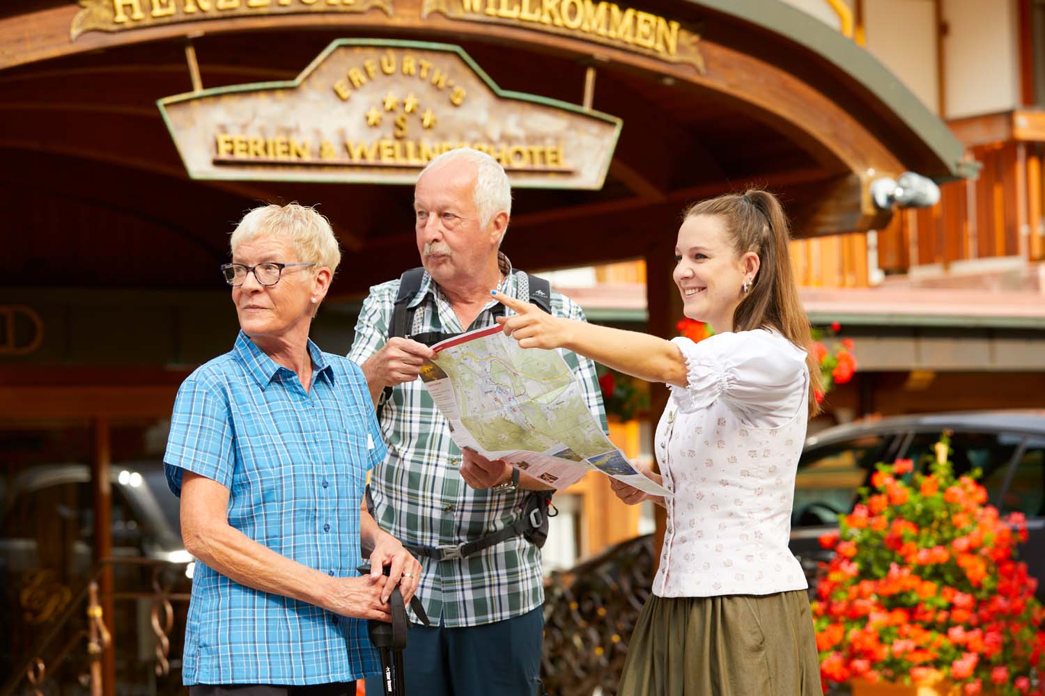 Eine junge Frau in traditioneller Kleidung hilft zwei älteren Erwachsenen mit einer Landkarte vor einem Hotel, während im Hintergrund Blumen und ein Willkommensschild zu sehen sind.