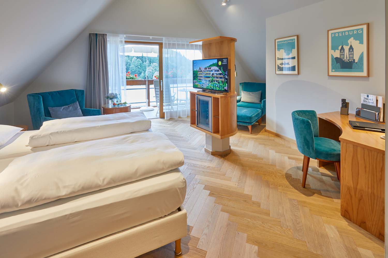 Ein helles Hotelzimmer mit einem Doppelbett, einem Fernseher auf einem Holzständer, einem Schreibtisch mit Stuhl, zwei tealfarbenen Sesseln, gerahmter Wandkunst und einem Balkon mit Sitzgelegenheiten im Freien mit Blick ins Grüne.