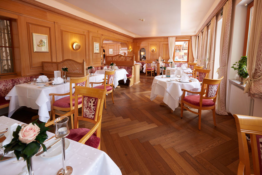 Elegantes Restaurant mit Holzböden und getäfelten Wänden, mit runden, weiß gedeckten Tischen, ordentlich aufgestellten Stühlen und Blumenschmuck, beleuchtet von natürlichem Licht aus großen Fenstern.