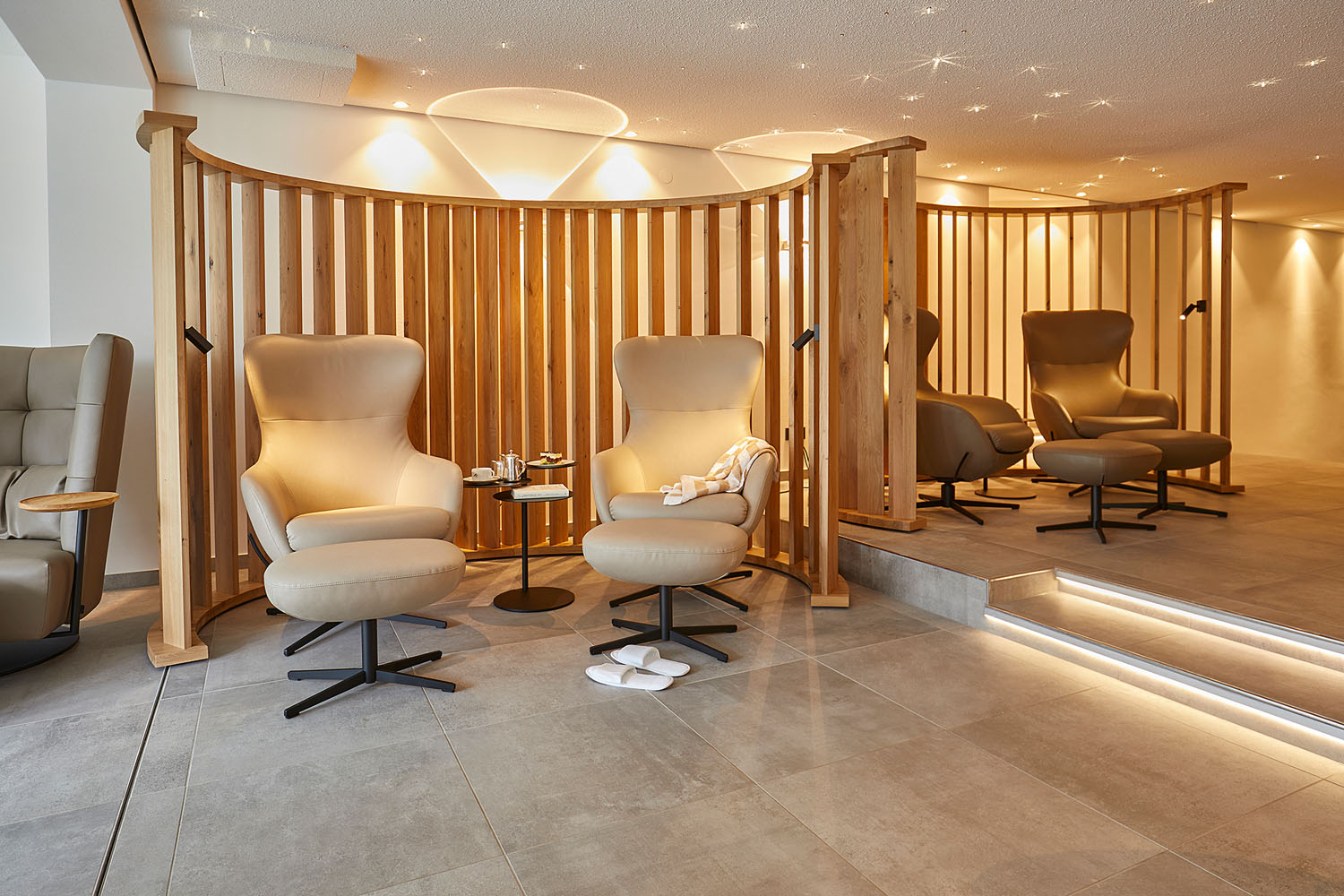 Zwei moderne beigefarbene Loungesessel mit passenden Fußstützen stehen neben einem kleinen Tisch mit Teetassen und Handtüchern. Hinter den Sesseln wölben sich Trennwände aus Holzlatten. Eine sanfte Beleuchtung schafft eine entspannende, spa-ähnliche Atmosphäre.