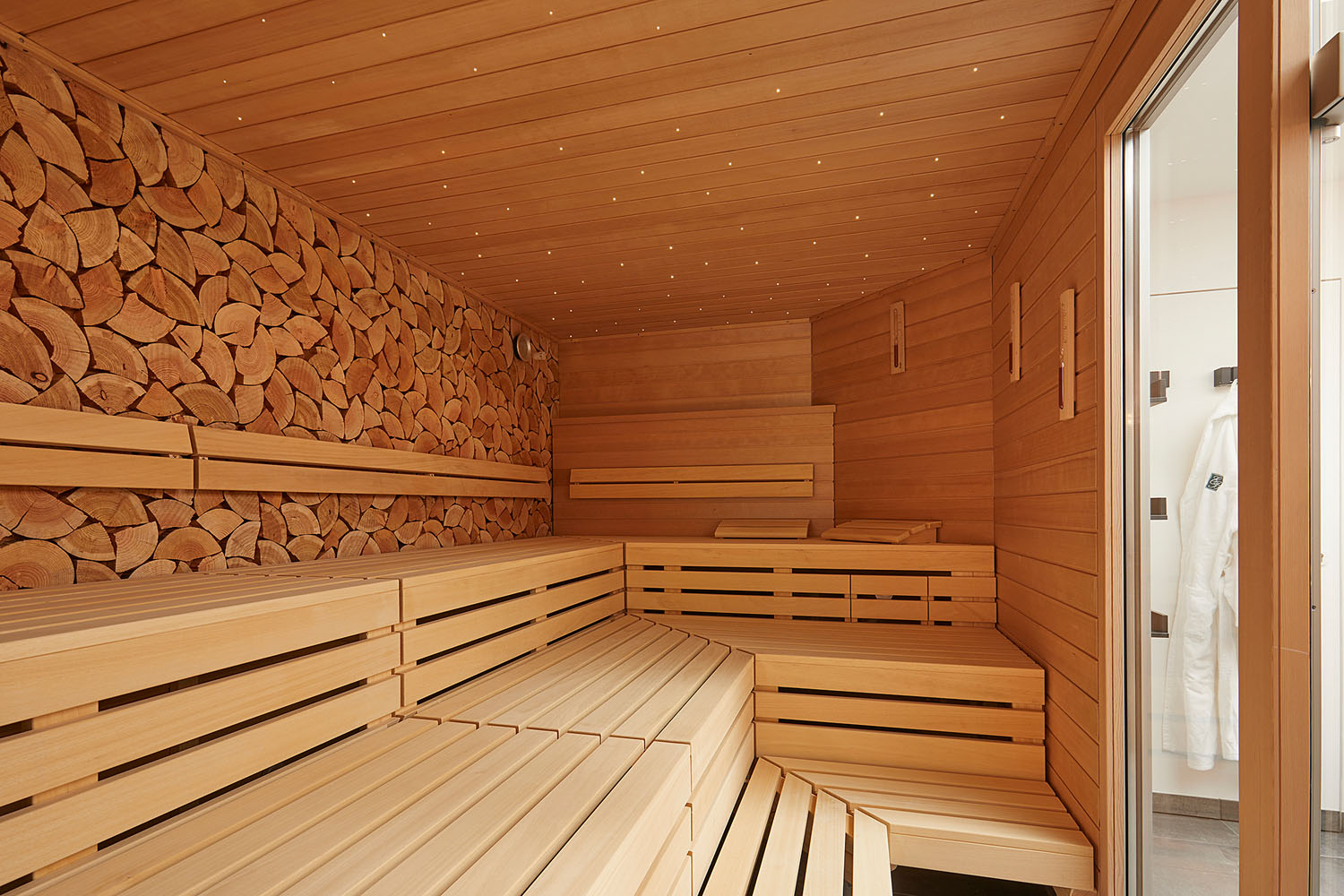 Eine moderne Holzsauna mit gestuften Bänken, ordentlich gestapeltem Brennholz an einer Wand und einer Glastür, vor der weiße Bademäntel hängen.