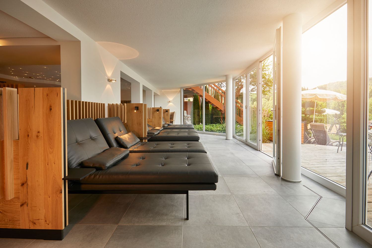 Eine moderne Entspannungslounge mit schwarzen Ledersesseln, hölzernen Trennwänden und großen, raumhohen Fenstern, die das Sonnenlicht hereinlassen und den Blick auf eine Außenterrasse mit Sonnenschirmen und Grünflächen freigeben.
