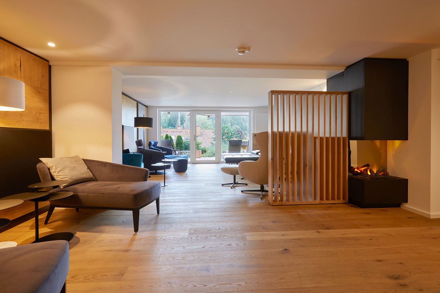 Ein modernes Wohnzimmer mit Holzböden, modernen Möbeln, einem Paravent und einem Kamin. Große Glastüren an der Rückseite öffnen sich zu einer Terrasse mit Blick auf Bäume und Grünflächen draußen.