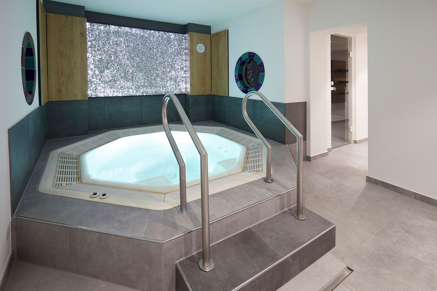 Ein moderner Indoor-Whirlpool mit Metallgeländern, umgeben von grauen Fliesen, vor einer dekorativen Wand mit funkelnden Mosaikfliesen und runden Spiegeln auf jeder Seite. Der Raum ist hell und sauber mit einem minimalistischen Design.