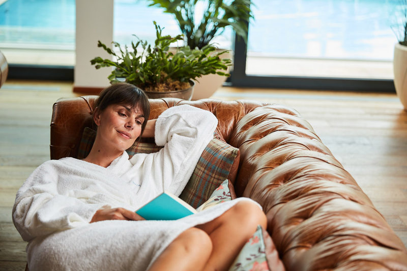 Eine Frau in einem weißen Bademantel entspannt sich auf einem braunen Ledersofa und umarmt Wellness für Frauen, während sie ein Buch liest. Sie lehnt sich zurück und stützt ihren Kopf auf ihre Hand, in der Nähe eines Fensters mit Pflanzen und einem ruhigen Blick auf den Pool draußen.