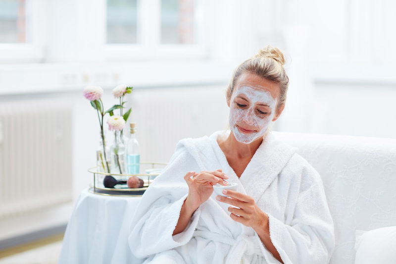 Eine Frau in einem weißen Bademantel sitzt auf einem Sofa und lächelt, während sie eine Gesichtsmaske aufträgt - Wellness für Frauen. Sie hält ein kleines Gefäß neben einem Beistelltisch mit Blumen und Schönheitsprodukten in ihrem hellen, luftigen Raum.