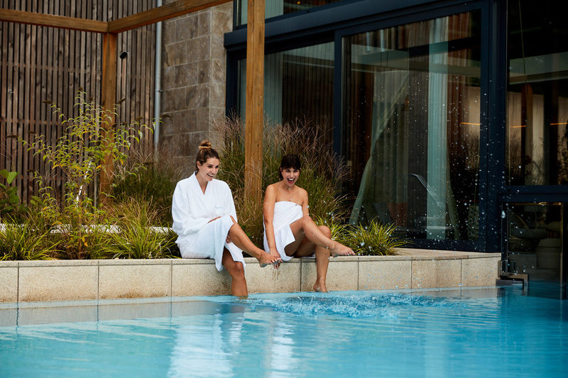 Zwei Frauen in Bademänteln sitzen am Rande eines Außenpools, lächeln und tauchen ihre Füße ins Wasser. Umgeben von üppigen Pflanzen und hohen Glasfenstern genießen sie einen Moment von Wellness für Frauen.