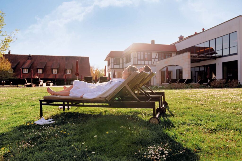Zwei Personen in weißen Bademänteln entspannen sich auf Liegestühlen auf einer Wiese vor einem modernen Gebäude mit großen Fenstern unter einem klaren blauen Himmel. Neben einem der Stühle stehen weiße Hausschuhe.
