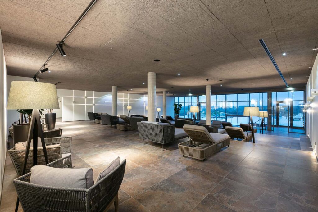 Ein geräumiges, modernes Wohnzimmer mit Korbstühlen und Liegestühlen, sanfter Beleuchtung durch Stehlampen, einem großen Fliesenboden und bodentiefen Fenstern, die den Blick nach draußen freigeben. Der Raum strahlt eine ruhige und einladende Atmosphäre aus.