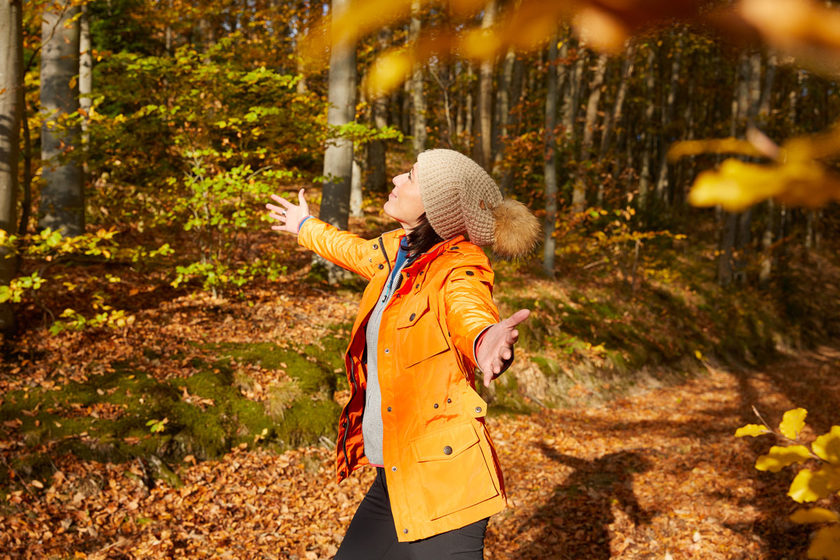 Eine Person in einer leuchtend orangefarbenen Jacke und einer beigen Strickmütze steht in einem sonnendurchfluteten Herbstwald mit ausgestreckten Armen, umgeben von bunten Herbstblättern und Bäumen.