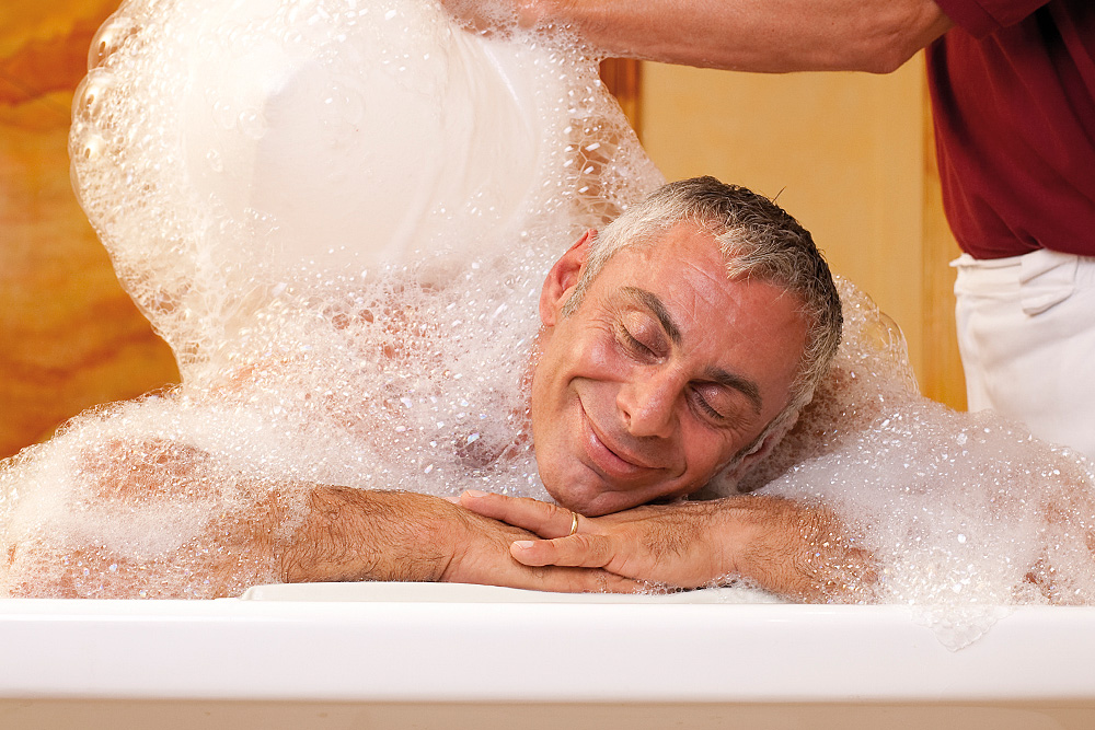 Ein Mann mit grauem Haar entspannt sich und lächelt in einer mit Seifenblasen gefüllten Badewanne, während jemand weitere Seifenblasen aus einem weißen Behälter über ihn schüttet.