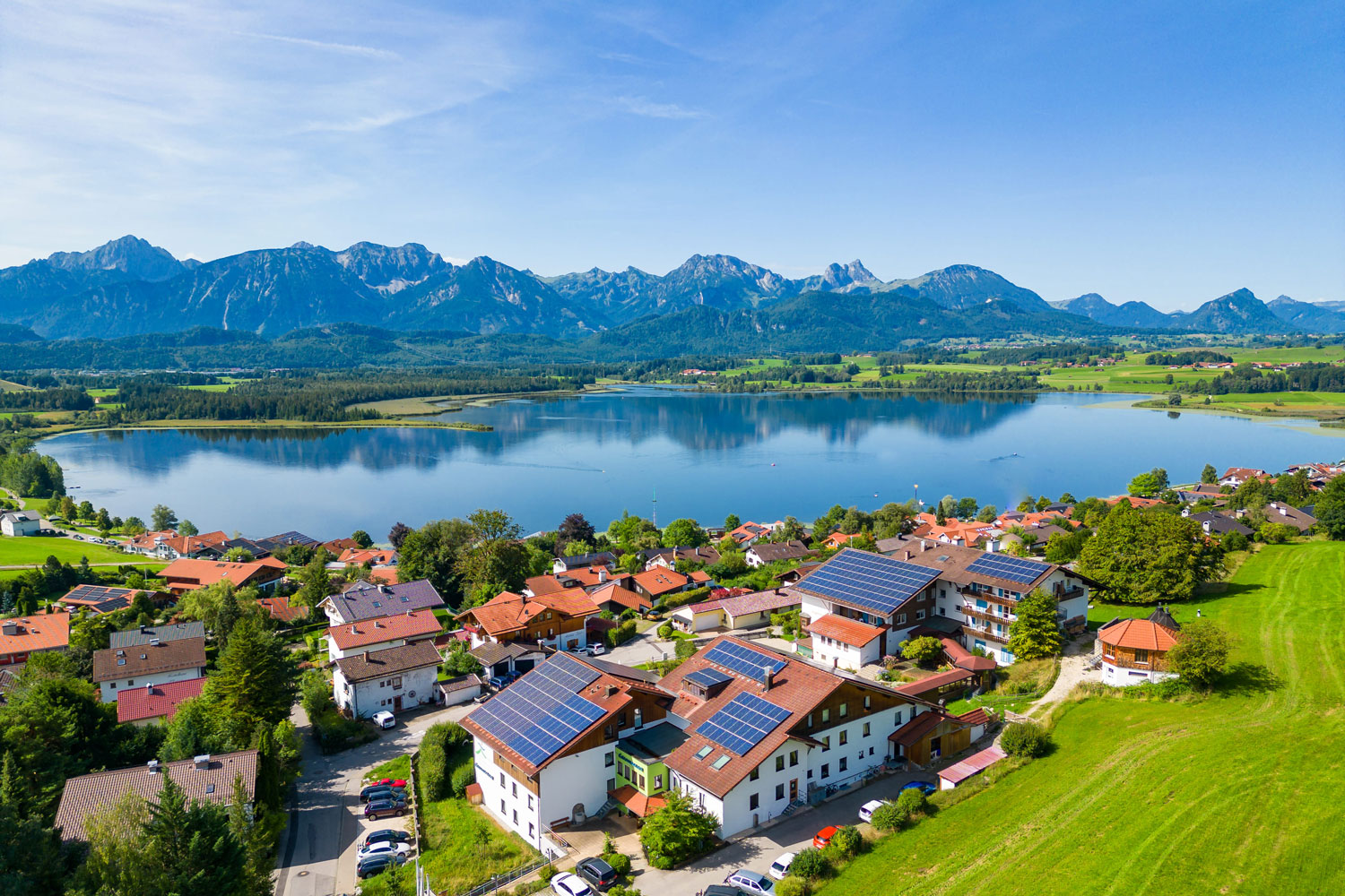 Luftaufnahme eines Seedorfs mit roten Dächern, Sonnenkollektoren und grünen Feldern - das Bio- & Wellnesshotel Eggensberger - an einem klaren blauen See und umgeben von Bergen unter einem strahlend blauen Himmel.