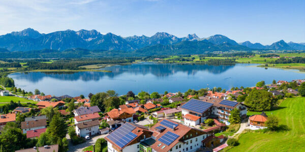 Luftaufnahme eines Dorfes mit roten Dächern und Sonnenkollektoren, ideal für Wandern und Wellness, an einem großen See mit grünen Feldern und hohen Bergen im Hintergrund unter einem klaren blauen Himmel.